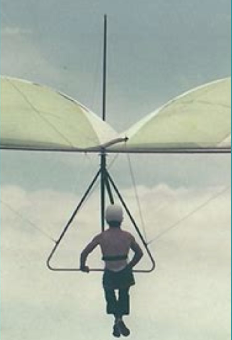 Early hang glider pilot seen from behind, hanging from the control bar against a grey sky
